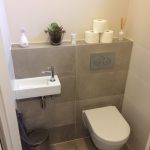 Compact toilet met strakke beige tegels, moderne inbouwspoeling, en een kleine wastafel met minimalistische accessoires.