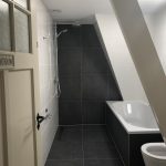 Badkamer in een zolderruimte met grijze wand- en vloertegels, een ligbad en inloopdouche, met een schuine wand en authentieke deur.