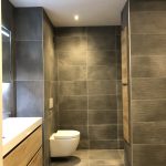 Moderne badkamer met elegante grijze wand- en vloertegels, zwevend toilet en een houten wastafelmeubel voor een minimalistische uitstraling.