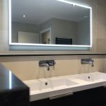 Ruime badkamer met dubbele wastafels, een verlichte spiegel en moderne kranen, afgewerkt met strakke wandtegels.