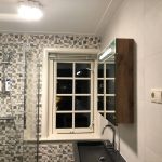 Badkamer met mozaïek wandtegels, houten spiegelkast, en een groot raam met klassieke roedeverdeling.