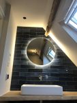 Stijlvolle badkamer met blauwe wandtegels, een grote ronde spiegel met sfeerverlichting en een houten wastafelmeubel, omgeven door stucwerk.