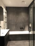 Elegante badkamer met grote grijze tegels, een inloopdouche met glazen wand, een vrijstaand bad en een strak wastafelmeubel.