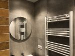 Badkamer met ronde spiegel, witte handdoekradiator en grijze betonlook wandtegels, met een moderne toiletpot en strakke afwerking.
