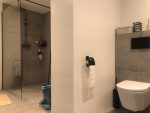 Ruime, luxe badkamer met een grote inloopdouche, modern toilet en een stijlvol ligbad, alles afgewerkt met grijze tegels en strakke lijnen.