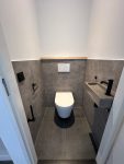 Modern toilet met strakke grijze tegels, zwarte wateraansluiting en minimalistisch design met houten accenten.